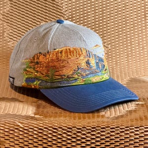 May include: A light gray baseball cap featuring a detailed embroidered landscape of orange rock formations, green vegetation, and a blue river. The cap has a solid blue brim, a blue top button, and a small black rectangular tag on the side.