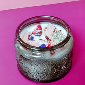 May include: A round, dark glass candle jar with an embossed sun pattern on the exterior. The white wax is topped with dried red rose petals, small blue stones, light pink crystals, and a silver-toned metal charm featuring the Libra scales symbol.