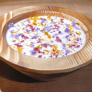 Puede incluir: Una vela decorativa en un cuenco de madera. La cera blanca est&aacute; adornada con p&eacute;talos de flores de colores en tonos morado, rojo y amarillo. Una sola mecha se encuentra en el centro de la vela.