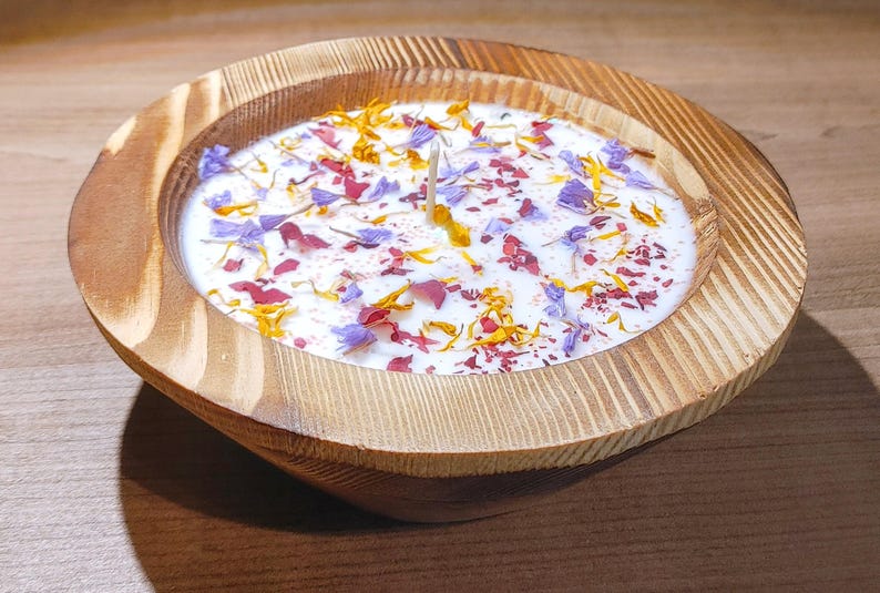 Puede incluir: Una vela decorativa en un cuenco de madera. La cera blanca est&aacute; adornada con p&eacute;talos de flores de colores en tonos morado, rojo y amarillo. Una sola mecha se encuentra en el centro de la vela.