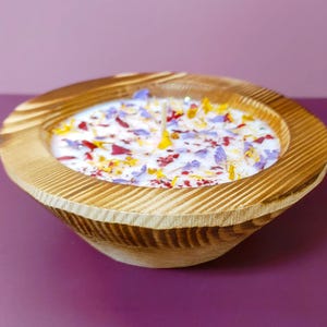 Puede incluir: Una vela en forma de cuenco de madera con una base de cera blanca, adornada con p&eacute;talos de flores de colores. El cuenco tiene un acabado de vetas de madera natural y est&aacute; sobre un fondo p&uacute;rpura. La vela tiene varias mechas.