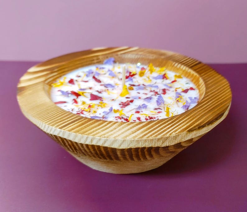 Puede incluir: Una vela en forma de cuenco de madera con una base de cera blanca, adornada con p&eacute;talos de flores de colores. El cuenco tiene un acabado de vetas de madera natural y est&aacute; sobre un fondo p&uacute;rpura. La vela tiene varias mechas.