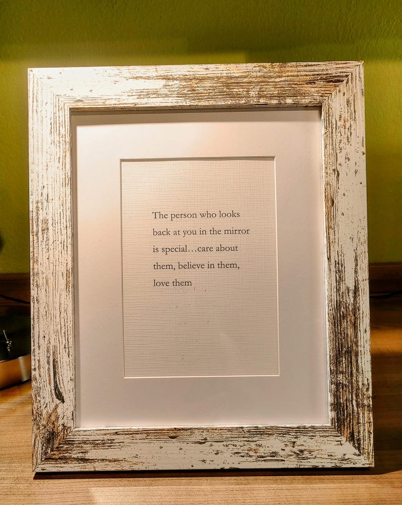 Op de afbeelding: Een ingelijste print met een witte passe-partout en een verweerde wit en bruin houten lijst. De print bevat de tekst: "De persoon die in de spiegel naar je kijkt is speciaal... zorg voor ze, geloof in ze, houd van ze."