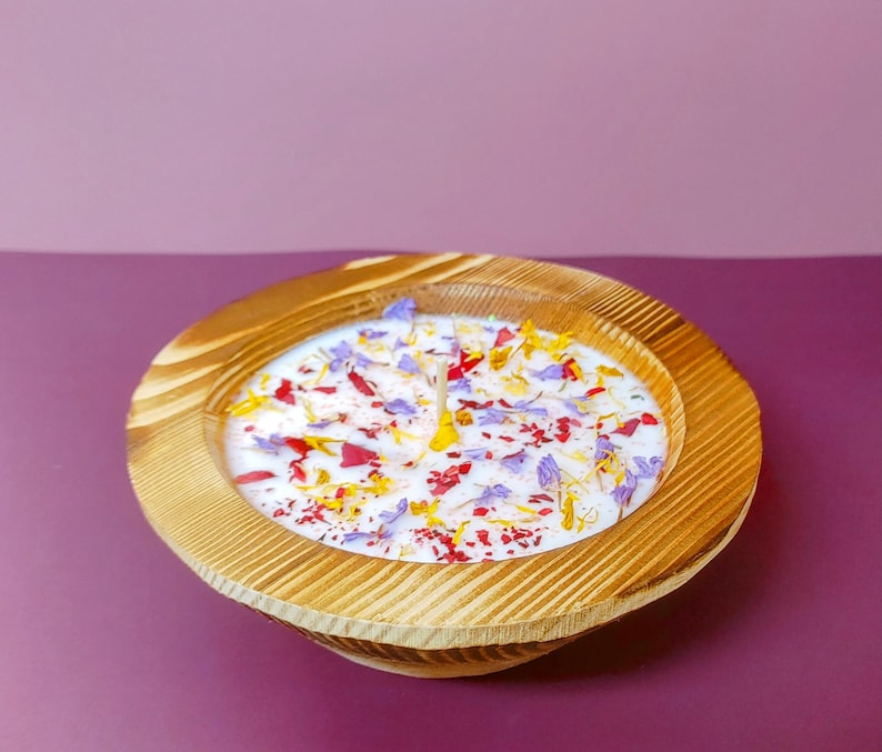 May include: A wooden bowl candle with a white wax base and embedded dried flowers in shades of purple, red, and yellow. The bowl is made of light brown wood with a natural wood grain pattern.