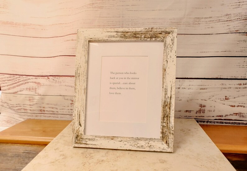 Op de afbeelding: Een ingelijste print met een wit, verweerd houten frame. De print heeft een witte passe-partout en een lichtroze achtergrond met de tekst: "De persoon die in de spiegel naar je kijkt is speciaal... zorg voor hen, geloof in hen, houd van hen."