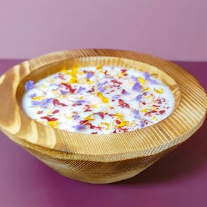 Puede incluir: Una vela en forma de cuenco de madera con una superficie de cera blanca, adornada con p&eacute;talos de flores de colores. El cuenco tiene un acabado de vetas de madera natural y se apoya sobre una superficie p&uacute;rpura. Es un art&iacute;culo decorativo.
