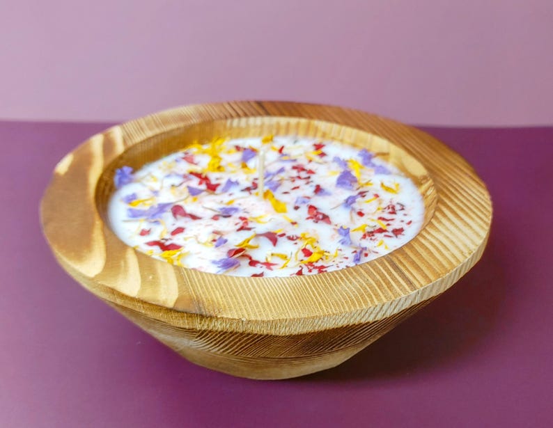 Puede incluir: Una vela en forma de cuenco de madera con una superficie de cera blanca, adornada con p&eacute;talos de flores de colores. El cuenco tiene un acabado de vetas de madera natural y se apoya sobre una superficie p&uacute;rpura. Es un art&iacute;culo decorativo.