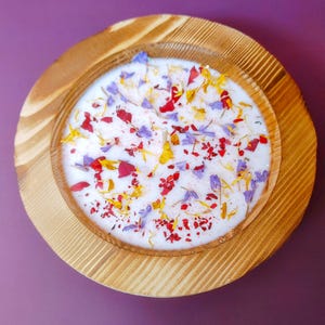 Puede incluir: Un portavelas redondo de madera con una vela blanca. La vela est&aacute; decorada con p&eacute;talos de flores coloridos en rojo, amarillo y morado. El portavelas de madera tiene un acabado de vetas naturales y est&aacute; sobre una superficie p&uacute;rpura.