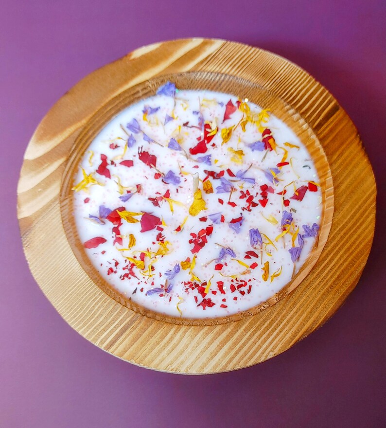 Puede incluir: Un portavelas redondo de madera con una vela blanca. La vela est&aacute; decorada con p&eacute;talos de flores coloridos en rojo, amarillo y morado. El portavelas de madera tiene un acabado de vetas naturales y est&aacute; sobre una superficie p&uacute;rpura.
