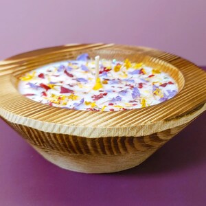 Puede incluir: Una vela en forma de cuenco de madera con una base de cera de color claro. La cera est&aacute; decorada con p&eacute;talos de flores de colores. El cuenco tiene un acabado de vetas de madera natural y un exterior acanalado. Se ven dos mechas.