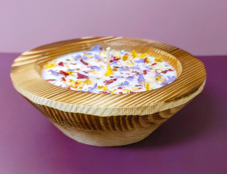 Puede incluir: Una vela en forma de cuenco de madera con una base de cera de color claro. La cera est&aacute; decorada con p&eacute;talos de flores de colores. El cuenco tiene un acabado de vetas de madera natural y un exterior acanalado. Se ven dos mechas.