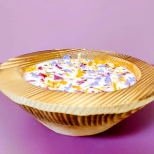 Puede incluir: Una vela en cuenco de madera con base de cera blanca, adornada con p&eacute;talos de flores de colores. El cuenco tiene un acabado de vetas de madera natural y una sola mecha. La vela est&aacute; sobre una superficie p&uacute;rpura.