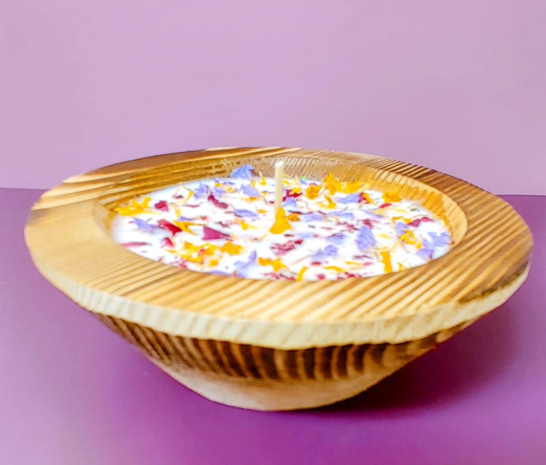 Puede incluir: Una vela en cuenco de madera con base de cera blanca, adornada con p&eacute;talos de flores de colores. El cuenco tiene un acabado de vetas de madera natural y una sola mecha. La vela est&aacute; sobre una superficie p&uacute;rpura.