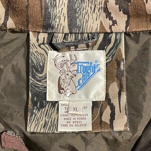 May include: Close-up of a camouflage jacket with a tag. The tag features a deer illustration and the words "Trophy Club". The jacket is brown, tan, and green. The tag also includes size information and material details.