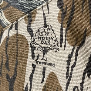 May include: Close-up of a camouflage pattern fabric with the "Mossy Oak" logo and "Treestand" text. The fabric features a mix of brown, tan, black, and green colors, creating a natural, woodsy appearance. The logo is a stylized tree.