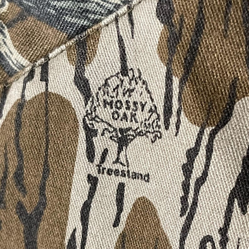 May include: Close-up of a camouflage pattern fabric with the "Mossy Oak" logo and "Treestand" text. The fabric features a mix of brown, tan, black, and green colors, creating a natural, woodsy appearance. The logo is a stylized tree.