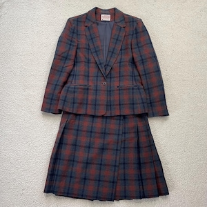 May include: A two-piece suit featuring a blazer and a skirt. The suit has a plaid pattern in shades of red, navy blue, and gray. The blazer has a single-button closure and notched lapels. The skirt is pleated.
