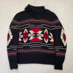May include: Black knit sweater with a geometric pattern in red, white, and gray. The sweater has a mock neck collar with three buttons and ribbed cuffs and hem. The design features diamond and zigzag patterns.