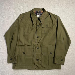 Puede incluir: Chaqueta verde oliva con cuatro bolsillos delanteros y cierres de botones. La chaqueta tiene un diseño acolchado en la parte superior del pecho y un forro a cuadros visible en el cuello. La etiqueta dice "SQUALITEX".