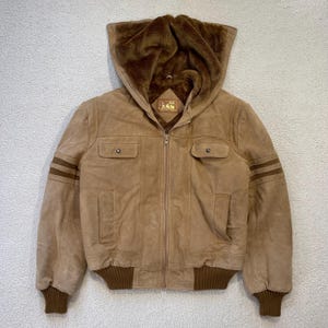 May include: A tan suede jacket with a hood lined with brown faux fur. The jacket features a zipper closure, two front pockets, and ribbed cuffs and hem. Brown stripes accent the left sleeve.