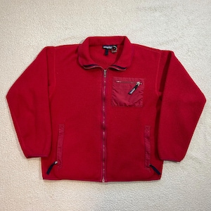Könnte beinhalten: Rote Fleecejacke mit durchgehendem Reißverschluss, einer kleinen Brusttasche und zwei Seitentaschen. Die Jacke hat einen Kragen und marineblaue Paspeln. Das Markenlogo ist am Kragen sichtbar. Ein warmes und bequemes Oberbekleidungsstück.