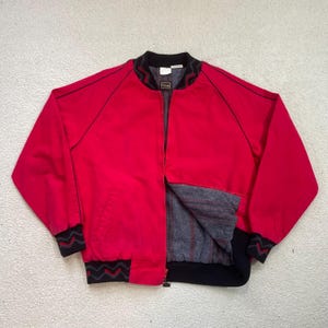 Könnte beinhalten: Eine leuchtend rote Bomberjacke mit schwarzen Verzierungen und einem durchgehenden Reißverschluss. Die Jacke hat schwarze Paspeln entlang der Nähte und ein gemustertes Design an den Manschetten und am Kragen. Das Innenfutter ist ein grau-rot gestreifter Stoff.