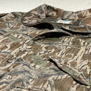 May include: Close-up of a camouflage jacket in shades of brown, tan, and green. The jacket features a button-up front, a collar, and two front pockets. The fabric has a textured appearance with a pattern resembling leaves and branches.
