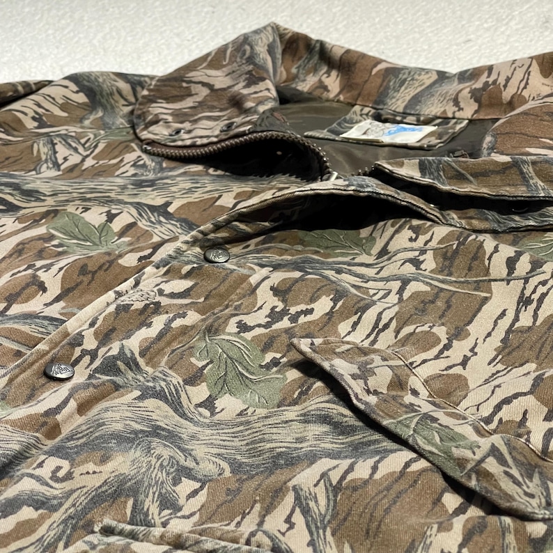 May include: Close-up of a camouflage jacket in shades of brown, tan, and green. The jacket features a button-up front, a collar, and two front pockets. The fabric has a textured appearance with a pattern resembling leaves and branches.