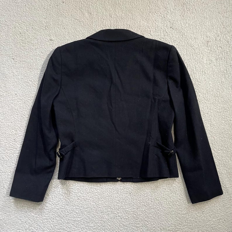May include: A black blazer with a notched lapel and long sleeves. The back of the jacket features a tailored design with decorative buttons and a fitted silhouette. The blazer is made of a smooth, dark fabric.