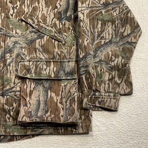 May include: Camouflage jacket with a brown, green, and tan tree bark pattern. The jacket has two large front pockets and a sleeve cuff. The fabric is a durable material, suitable for outdoor activities.