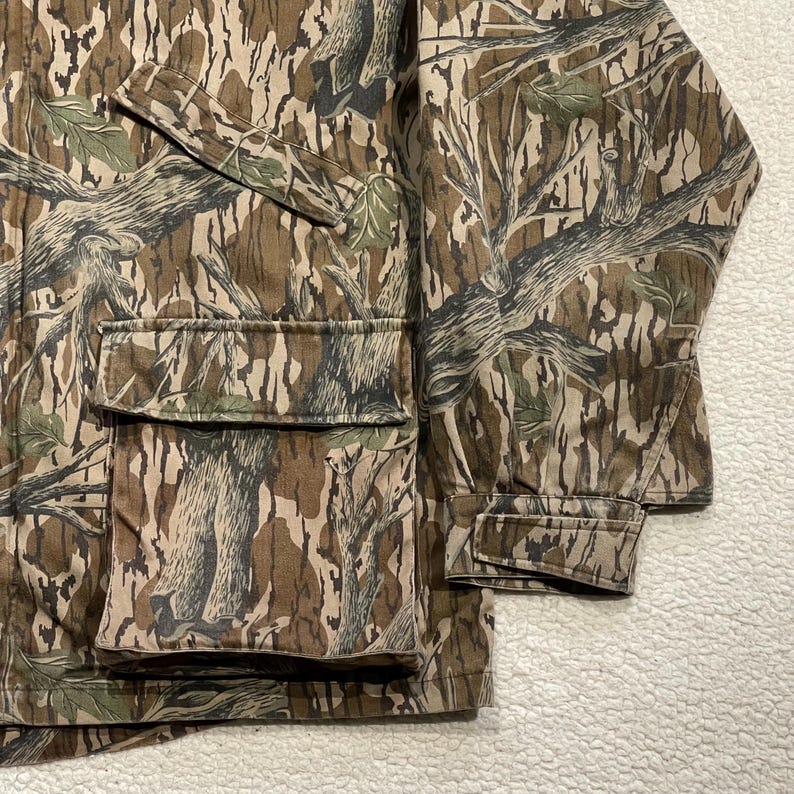 May include: Camouflage jacket with a brown, green, and tan tree bark pattern. The jacket has two large front pockets and a sleeve cuff. The fabric is a durable material, suitable for outdoor activities.