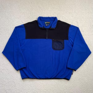 May include: A blue and black fleece pullover with a quarter-zip closure. The upper chest and collar are black, while the body is blue. It features a small mesh pocket on the chest and a small logo on the sleeve.
