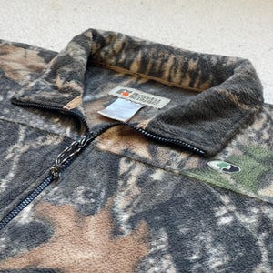 May include: A brown, black, and tan camouflage fleece jacket with a zipper closure. The jacket has a Russell Outdoors logo on the front.