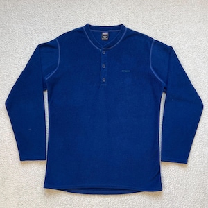 May include: A blue long-sleeve shirt with a button-up collar and a single button. The shirt has a small logo on the left chest.