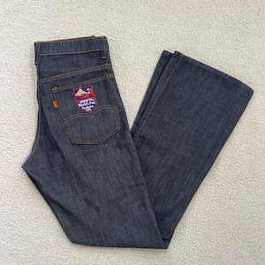 Może przedstawiać: Jeansy z ciemnego denimu z rozszerzaną nogawką. Tylna kieszeń posiada naszywkę z napisem "Expo '74 World's Fair Spokane USA" i grafiką gór. Jeansy mają klasyczny pięciokieszeniowy design oraz zapięcie na guzik i zamek błyskawiczny.