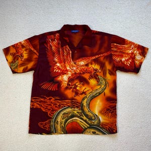 May include: A red short-sleeved button-up shirt with a graphic print of a golden snake and a red and orange eagle. The shirt has a blue button-up collar.