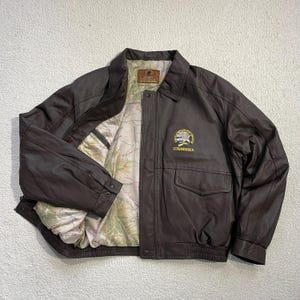 Könnte beinhalten: Braune Lederjacke mit Kartenmuster-Futter. Die Jacke hat einen Kragen, zwei Vordertaschen und ein "LIFE MEMBER"-Emblem, das in Gelb und Grün gestickt ist. Sie hat einen Reißverschluss und Knöpfe.