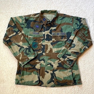 May include: Camouflage pattern military jacket with the text "BROWN" above the left pocket and "U.S. AIR FORCE" above the right pocket. The jacket has multiple pockets and patches, and is in shades of green, brown, and black.