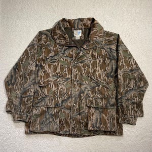Könnte beinhalten: Tarnmusterjacke mit durchgehendem Reißverschluss, zwei Vordertaschen und einem Kragen. Die Jacke hat ein braunes, grünes und beiges Farbschema, mit einer Knopfleiste und einem Logoaufnäher auf der Brust.