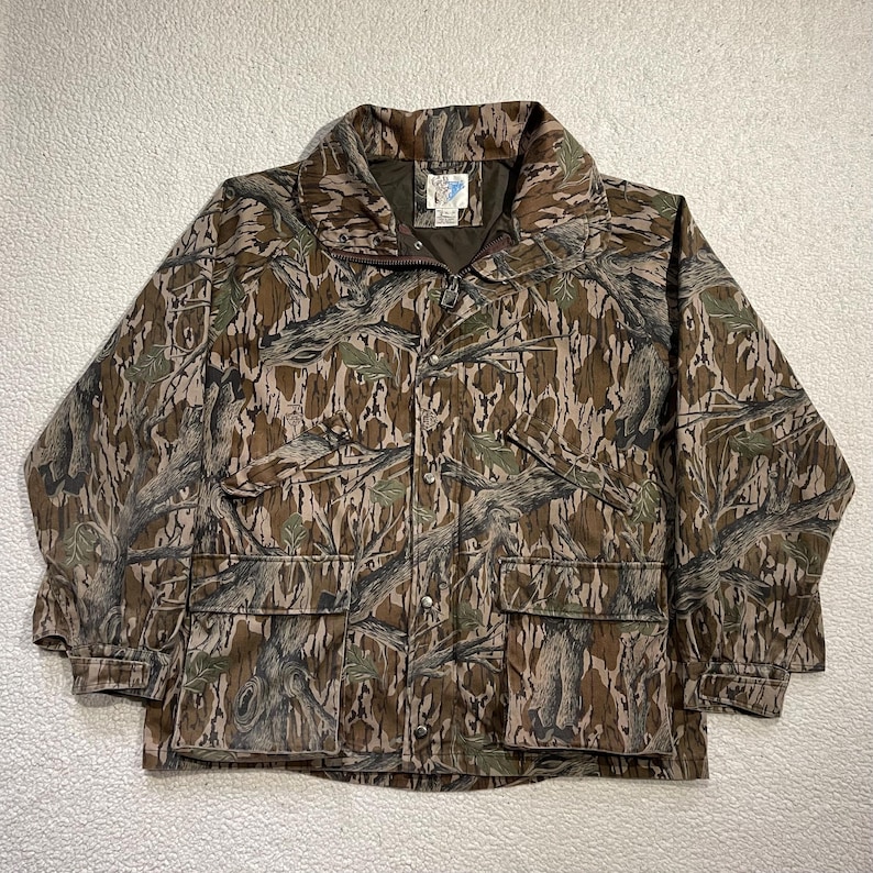 May include: Camouflage pattern jacket with a full zipper closure, two front pockets, and a collared neckline. The jacket features a brown, green, and tan color scheme, with a button-up front and a logo patch on the chest.