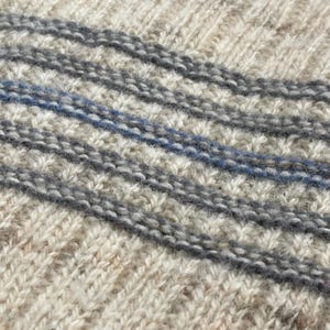 Pode incluir: Close-up de um tecido de malha com um padr&atilde;o texturizado. O su&eacute;ter apresenta listras diagonais em tons de cinza e uma &uacute;nica listra azul. A malha tem uma apar&ecirc;ncia quente e aconchegante, sugerindo um item feito &agrave; m&atilde;o ou artesanal.
