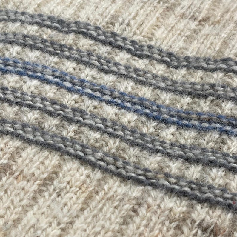 Pode incluir: Close-up de um tecido de malha com um padr&atilde;o texturizado. O su&eacute;ter apresenta listras diagonais em tons de cinza e uma &uacute;nica listra azul. A malha tem uma apar&ecirc;ncia quente e aconchegante, sugerindo um item feito &agrave; m&atilde;o ou artesanal.