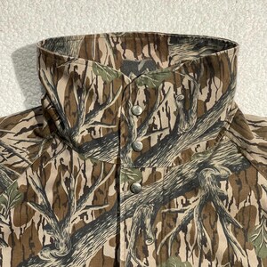 May include: A camouflage jacket with a high collar and three silver-colored buttons. The jacket features a pattern of brown, green, and tan tree branches and leaves. The collar is folded over and buttoned up.