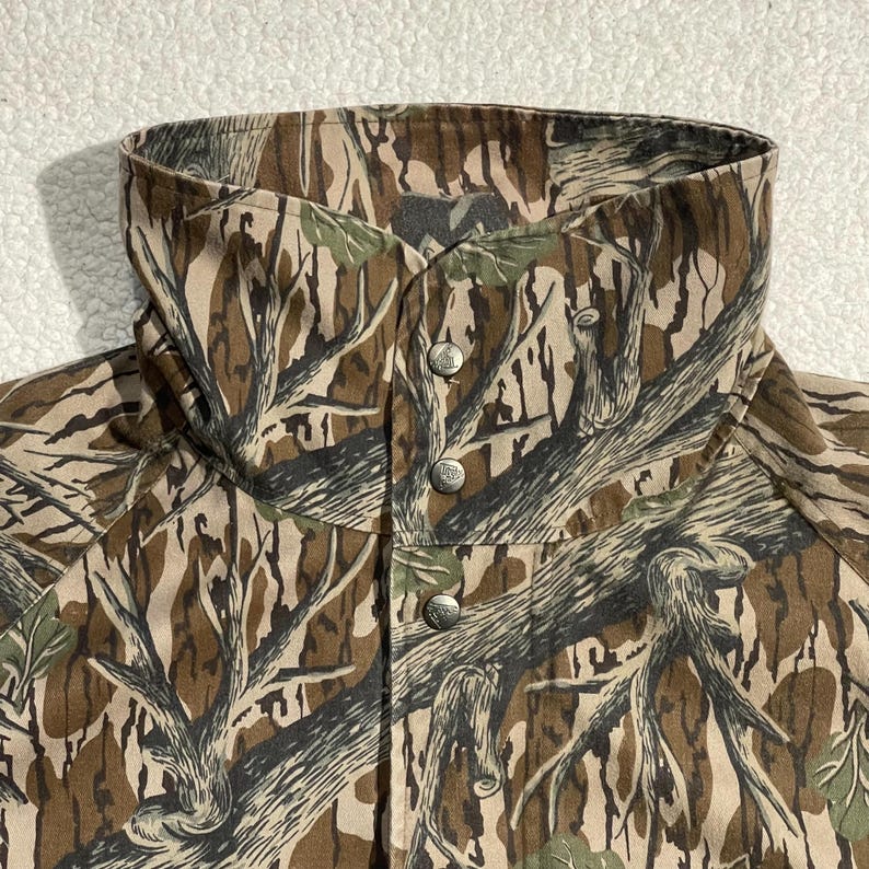 May include: A camouflage jacket with a high collar and three silver-colored buttons. The jacket features a pattern of brown, green, and tan tree branches and leaves. The collar is folded over and buttoned up.