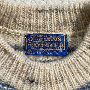 Pode incluir: Close-up de um su&eacute;ter de l&atilde; Pendleton bege e azul. O su&eacute;ter tem uma textura de malha canelada e uma etiqueta azul que diz "Pendleton" e "100% Virgin Wool". A etiqueta tamb&eacute;m diz "Made in U.S.A."