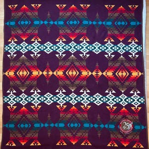 May include: A dark purple blanket with a geometric pattern in shades of red, orange, teal, brown, and white. The blanket features a repeating diamond and triangle design, with a circular emblem in the lower right corner.