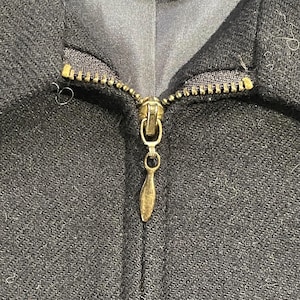 May include: Close-up of a black jacket zipper. The zipper is gold-colored and has a teardrop-shaped pull. The jacket fabric has a subtle textured pattern.