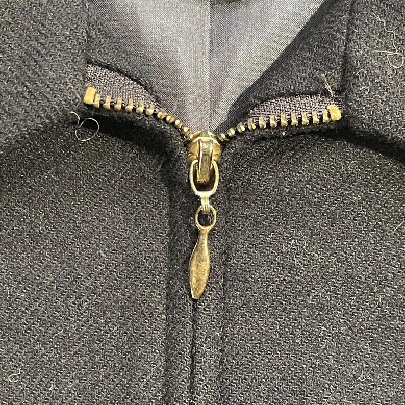 May include: Close-up of a black jacket zipper. The zipper is gold-colored and has a teardrop-shaped pull. The jacket fabric has a subtle textured pattern.