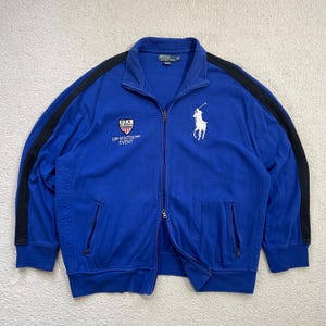 May include: A blue zip-up jacket with black stripes on the sleeves. Features a white polo player logo and text that reads "USA" and "Winter Event". The jacket has two front pockets and a ribbed hem and cuffs.