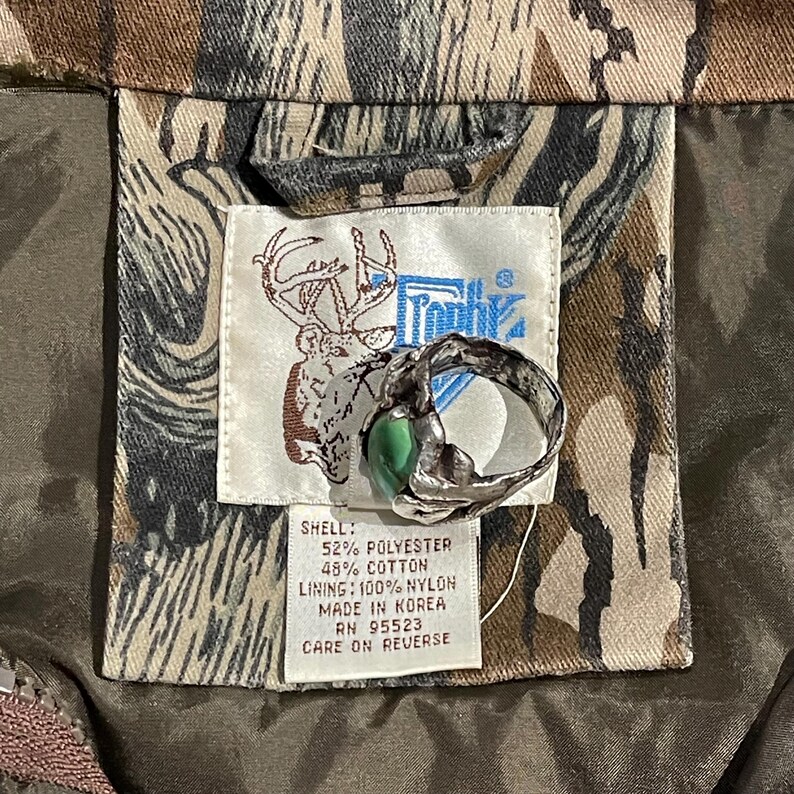 May include: A silver ring with a green stone is displayed on a camouflage jacket label. The label reads "Trophy" with a deer logo and fabric composition details: 52% polyester, 48% cotton, and 100% nylon lining.